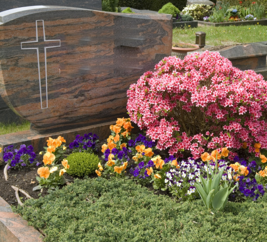 A gravestone with a cross engraving is surrounded by vibrant flowers, including pink blossoms, orange and purple pansies, in a well-kept cemetery.